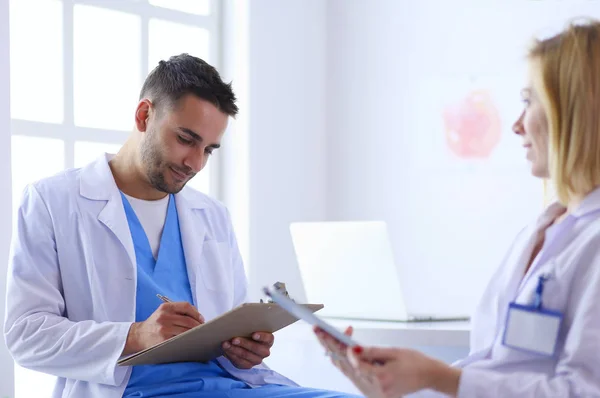 Schöner Arzt spricht mit junger Ärztin und macht sich Notizen, während er in seinem Büro sitzt. — Stockfoto