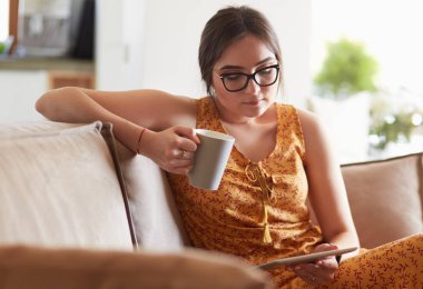 Genç kadın evinde kanepede kahve içerken tablet kullanıyor.