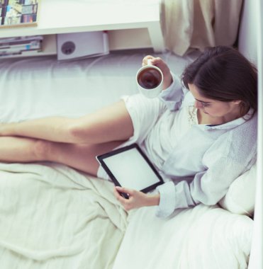 Sabahları yatakta otururken tablet kullanan ve kahve içen kadın portresi