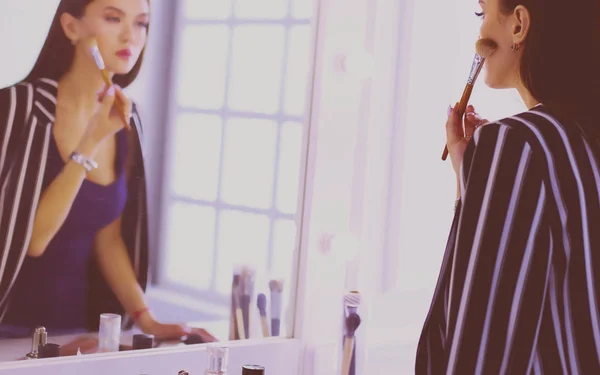 Красивая девушка смотрит в зеркало и наносит косметику большой кистью — стоковое фото