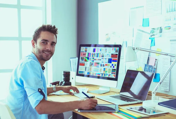 Retrato del joven diseñador sentado en el estudio gráfico frente a la computadora portátil y el ordenador mientras trabaja en línea. — Foto de Stock