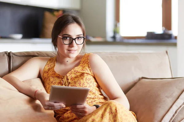 美丽的年轻黑发女人在家里坐在一张或settee使用她的平板电脑和微笑 — 图库照片