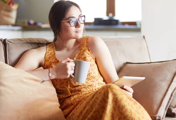 Mujer joven utiliza la tableta mientras se relaja en casa con café en el sofá sofá — Foto de Stock