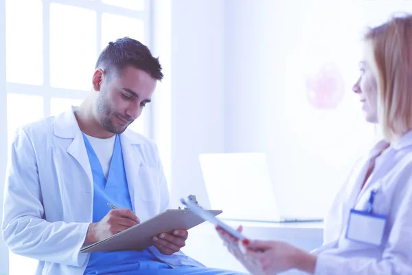 Médico bonito está conversando com jovem médica e fazendo anotações enquanto está sentado em seu escritório. — Fotografia de Stock