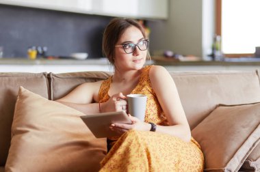 Genç kadın evinde kanepede kahve içerken tablet kullanıyor.