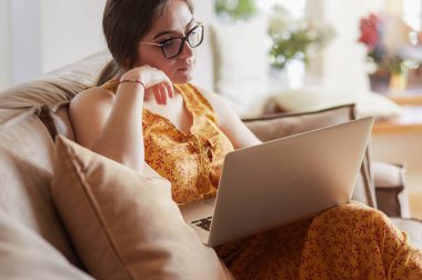 Evde dizüstü bilgisayar üzerinde çalışan kanepede oturan genç kadın