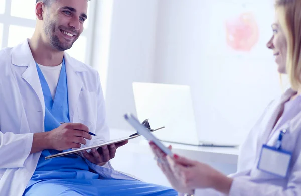 Médico bonito está conversando com jovem médica e fazendo anotações enquanto está sentado em seu escritório. — Fotografia de Stock