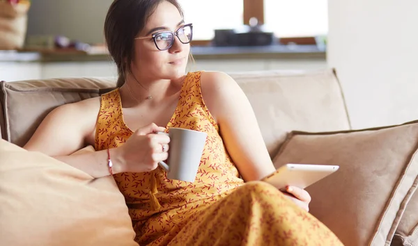 Young woman uses tablet while relaxing at home with coffee on the sofa couch — Stock Photo, Image
