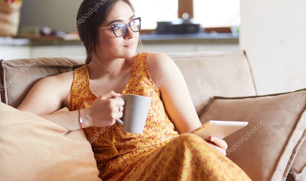 Young woman uses tablet while relaxing at home with coffee on the sofa couch