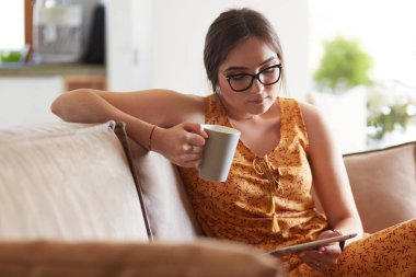Genç kadın evinde kanepede kahve içerken tablet kullanıyor.