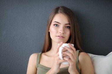 Güzel bir kadın beyaz bir kanepede bir fincan kahveyle dinleniyor.