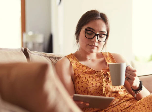 Giovane donna utilizza tablet mentre si rilassa a casa con il caffè sul divano — Foto Stock