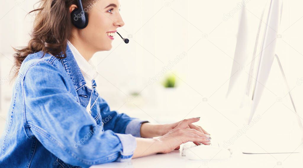 Happy charming young woman sitting and working with laptop using headset in office