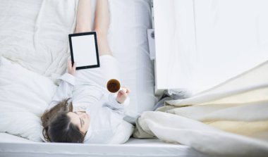 Sabahları yatakta otururken tablet kullanan ve kahve içen kadın portresi