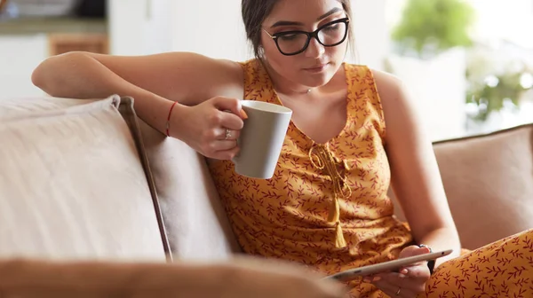 Genç kadın evinde kanepede kahve içerken tablet kullanıyor. — Stok fotoğraf