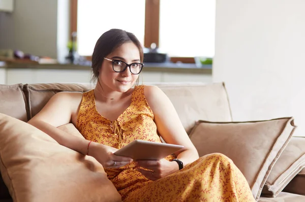 Piękna młoda kobieta brunetka w domu siedzi na kanapie lub settee za pomocą tabletu komputera i uśmiechnięty — Zdjęcie stockowe