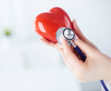 Steteskoplu bir doktor, beyaz arka planda kırmızı kalbi inceliyor.