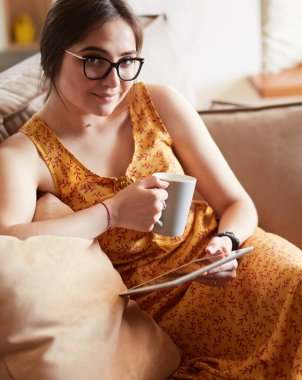 Genç kadın evinde kanepede kahve içerken tablet kullanıyor.