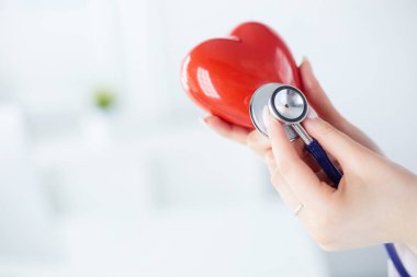 Steteskoplu bir doktor, beyaz arka planda kırmızı kalbi inceliyor.
