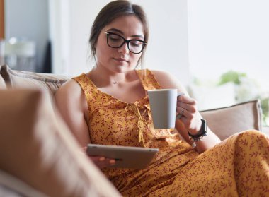 Genç kadın evinde kanepede kahve içerken tablet kullanıyor.