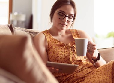 Genç kadın evinde kanepede kahve içerken tablet kullanıyor.