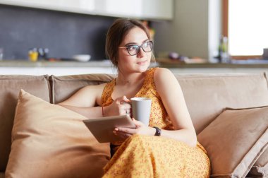 Genç kadın evinde kanepede kahve içerken tablet kullanıyor.