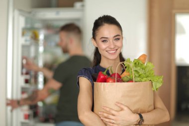 Mutfakta genç bir çift, alışveriş torbası taşıyan bir kadın.