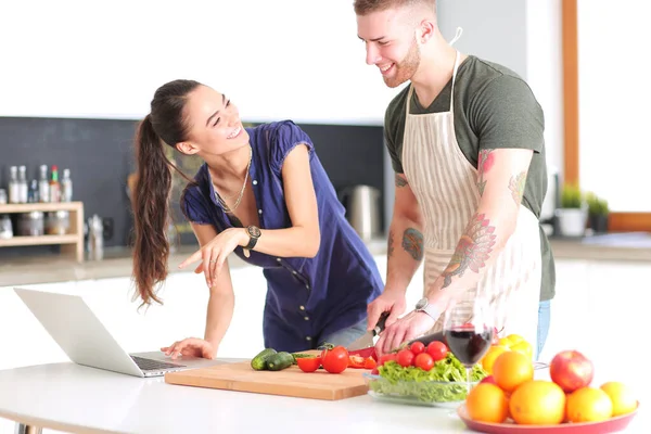 Hombre joven cortando verduras y mujer de pie con el ordenador portátil en la cocina — Foto de Stock