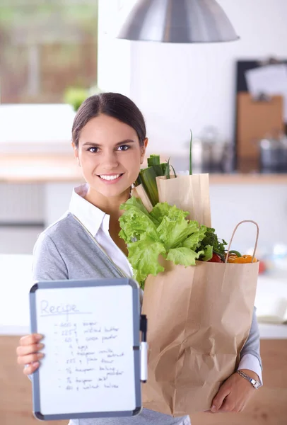 Mujer joven sosteniendo bolsa de la compra de comestibles con verduras. De pie en la cocina — Foto de Stock