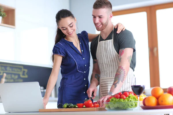 Jongeman snijden groenten en vrouw staan met laptop in de keuken — Stockfoto