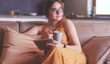 Genç kadın evinde kanepede kahve içerken tablet kullanıyor.