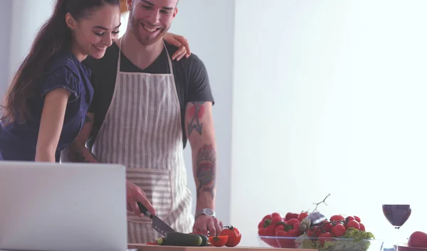 Młody mężczyzna kroi warzywa i kobieta stojąc z laptopem w kuchni — Zdjęcie stockowe