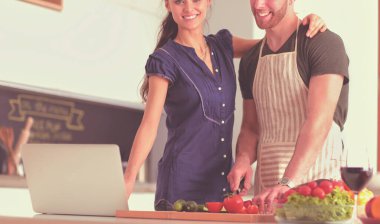 Sebze doğrayan genç bir adam ve mutfakta dizüstü bilgisayarı olan bir kadın.