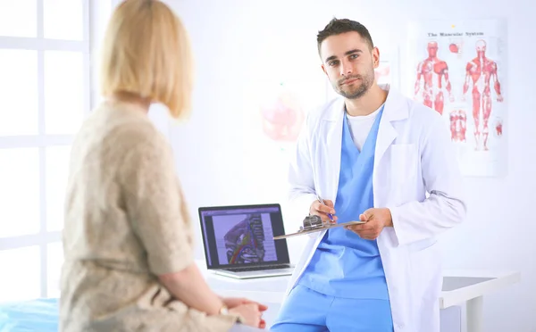 Beau médecin parle avec une jeune patiente et prend des notes alors qu'il est assis dans son bureau — Photo