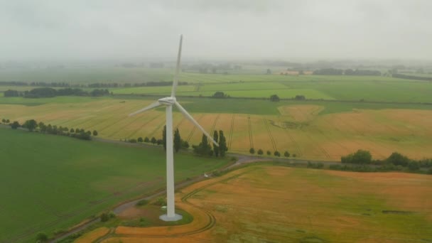 AERIAL: Vista del campo amarillo agrícola brumoso con turbinas de viento individuales que generan viento a través de la energía para un desarrollo sostenible en Alemania — Vídeo de stock