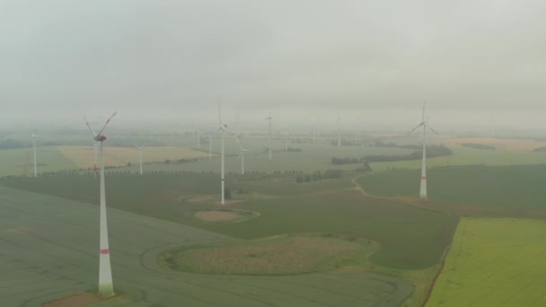 AERIAL: Zicht over mistig agrarisch geel veld met meerdere windturbines Energiestroom opwekken door wind voor een duurzame ontwikkeling in Duitsland — Stockvideo