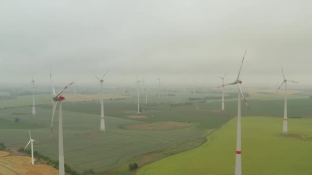 AERIAL: Meerdere windturbines op een rijk geel landbouwveld in mist die door de kracht van de wind draait en op een groene ecologische manier hernieuwbare energie opwekt voor de planeet — Stockvideo