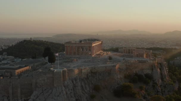 Vuelo aéreo hacia la Acrópolis de Atenas con bandera griega ondeando en la hermosa luz dorada del atardecer de la hora con el océano en la distancia — Vídeos de Stock