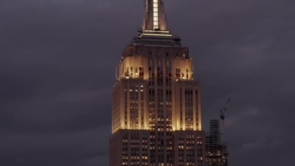 AERIAL: Voando até o Empire State edifício à noite em Manhattan cercado por arranha-céus em movimentada cidade — Vídeo de Stock