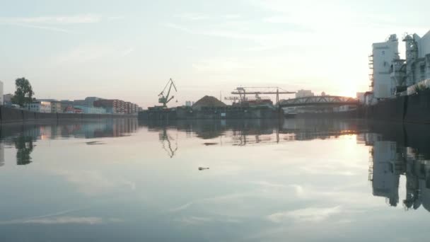 Una botella de vidrio vacía flotando en el río con área portuaria y contenedores en el fondo, vuelo aéreo bajo sobre el agua en los reflejos de la luz del sol de la madrugada — Vídeo de stock