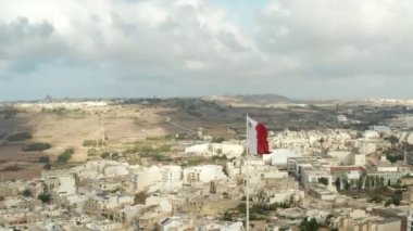 Rüzgârda dalgalanan Malta bayrağı Gozo Adası 'nın manzaralı havadan yakından sola kayıyor.