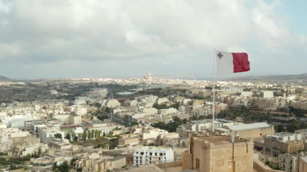 Slow Establishing Shot passing by Flag of Malta waving in revealing City on Gozo Island with cloudy Blue Sky, Aerial forward — стоковое видео