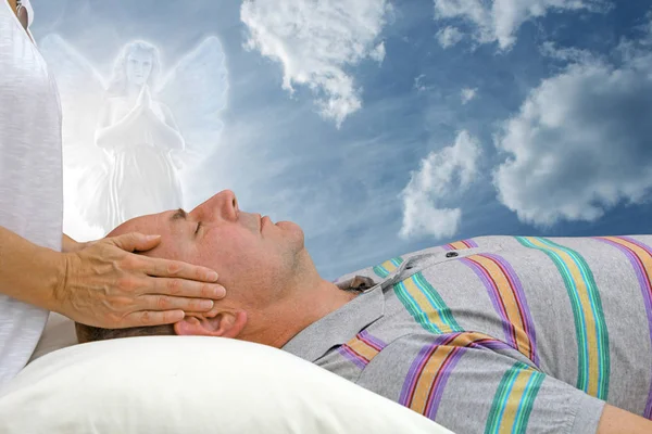 Out door energy healing with Guardian Angel - female with hands hovering over man's forehead, laid supine, against a beautiful sky with angel over looking and praying
