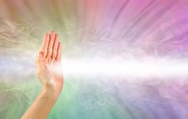 Pranic healer using right hand to beam healing energy -  female hand facing out with a beam of white energy streaming out against a rainbow coloured energy field background with copy space