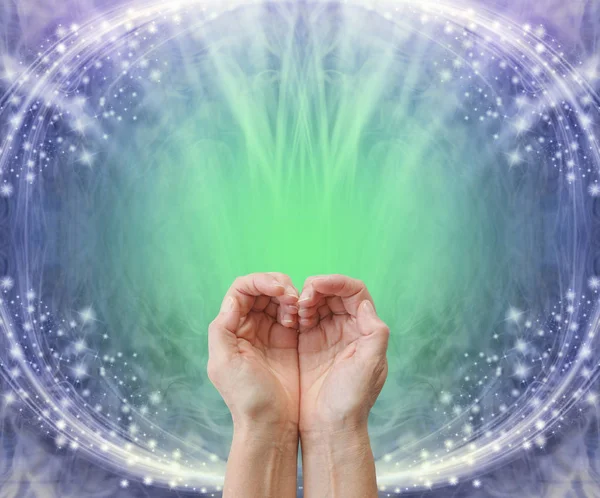 Sending heart chakra healing energy background - pair of cupped hands making a heart shape against a blue and green back ground with copy space and white sparkles creating an oval frame