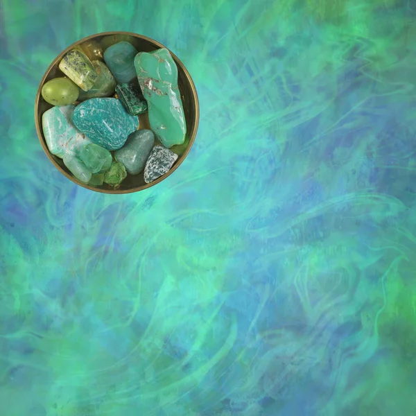 Green Heart Chakra Healing Crystal Selection - Circular brass dish with 10 different green coloured tumbled crystals against a wispy blue green ethereal background with copy space