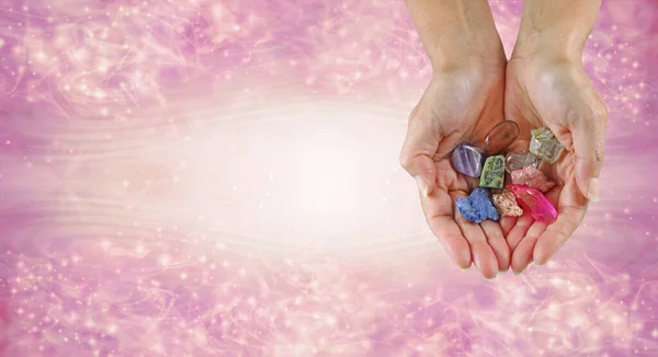 Crystal therapy message background banner - female hands cupped with a selection of raw and tumbled healing stones against a pink sparkling flowing background with space for text