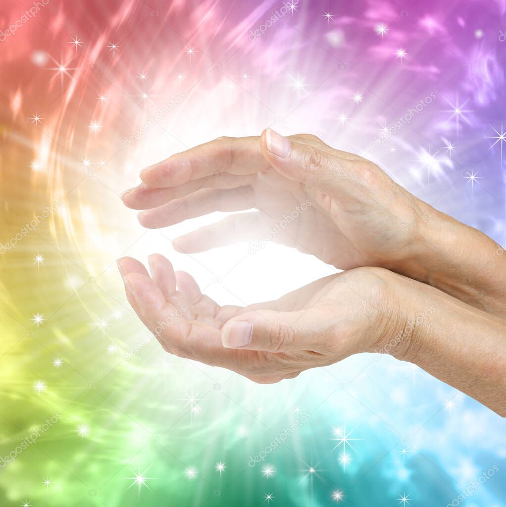 Colour Therapy Healing Hands Concept - female hands with bright white healing energy between against a rotating rainbow coloured energy formation background with copy space