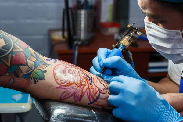 tattooist with his machine tattooing on the arm with the design drawn and using protective mask against covid 19, concept of art and design