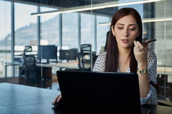 young, beautiful and empowered female entrepreneur, talking on a smartphone on speakerphone while working on her laptop in the office of meetings, business concept, technology and woman